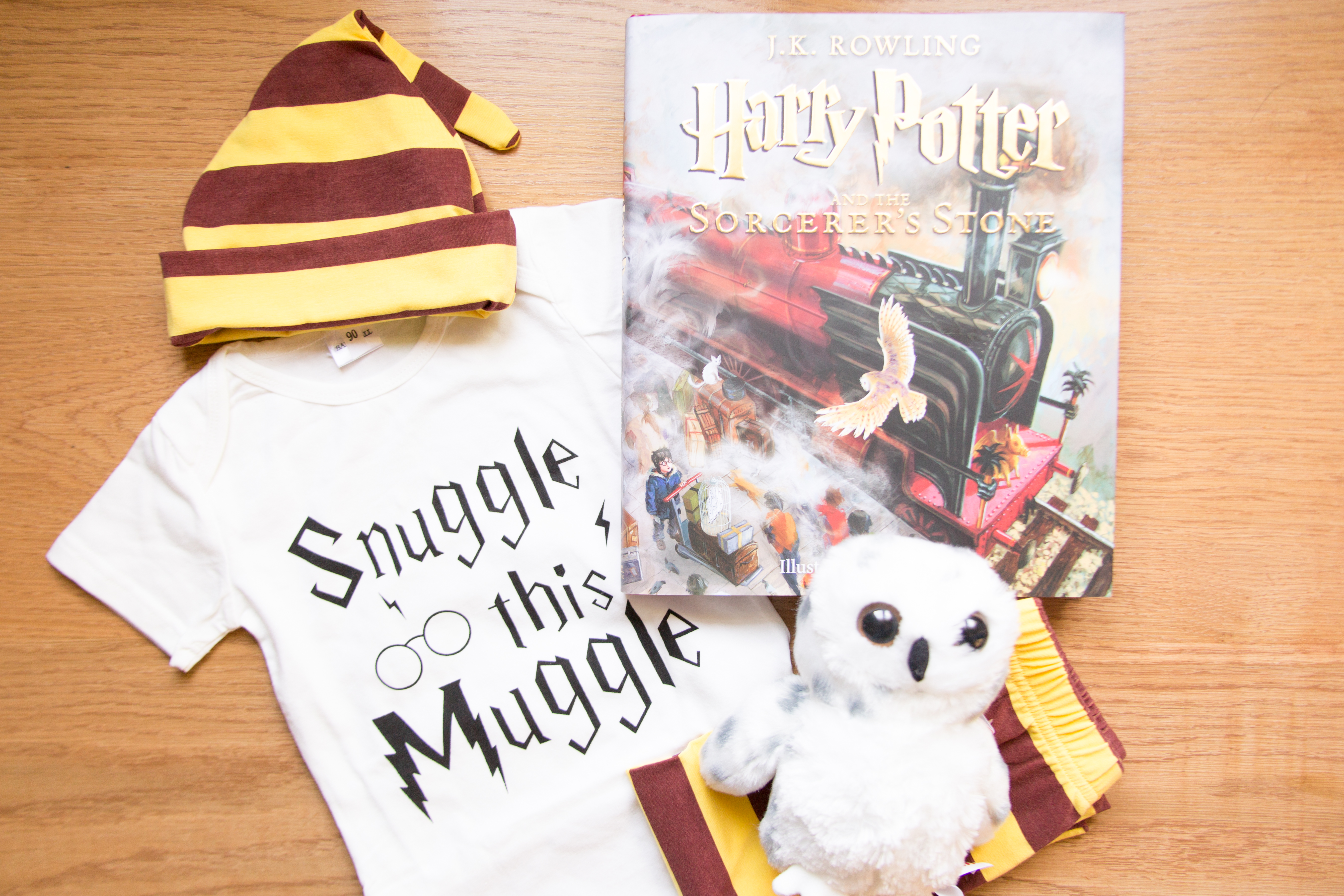 White baby clothes with the words snuggle this muggle with a red and yellow striped hat and pants on top with a copy of Harry Potter and the Sorcerers Stone to the right